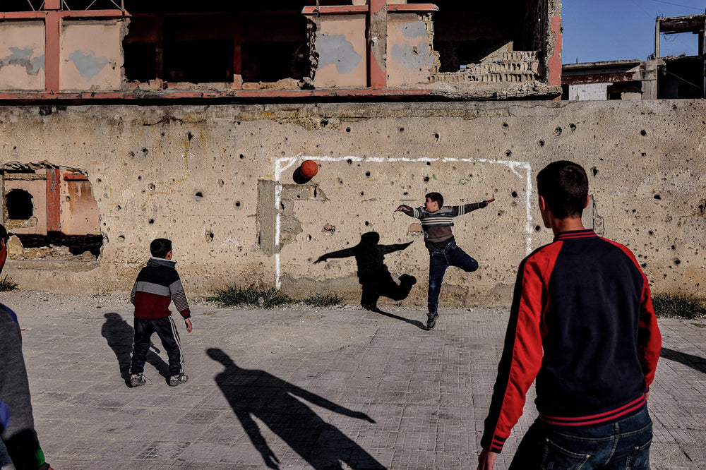 Children play football in front of a damaged wall, with one child leaping toward a red ball.