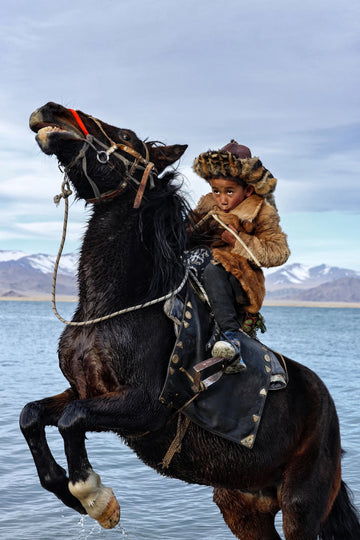 Young Kazakh eagle hunter riding through water in Mongolia — photograph by Laurin Strele