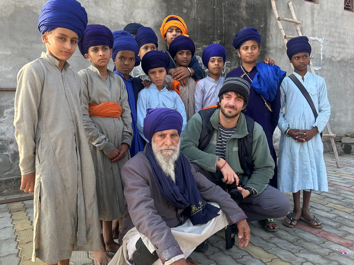 A photographer poses with a group in blue and orange turbans beside a grey wall.